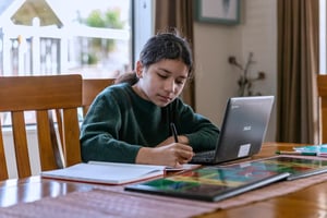 Student home learning taking notes in book while looking at chromebook
